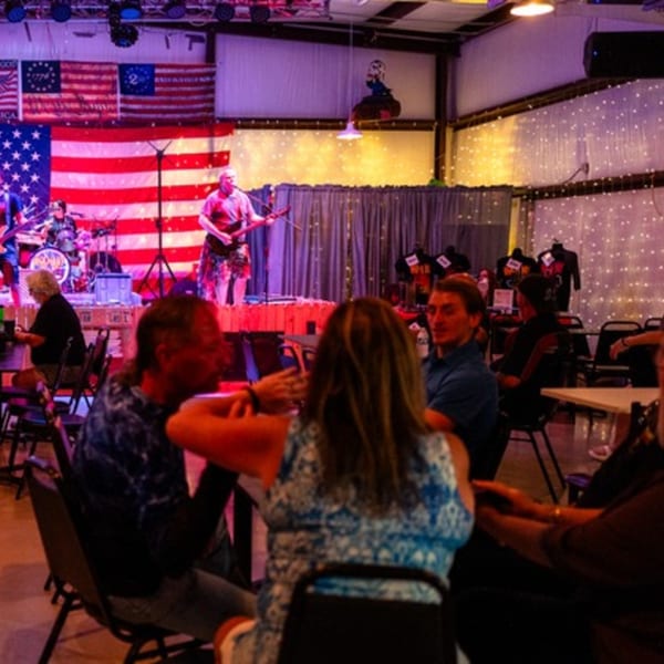 A band performs on stage with American flags behind them, while patrons sit at tables in the foreground.