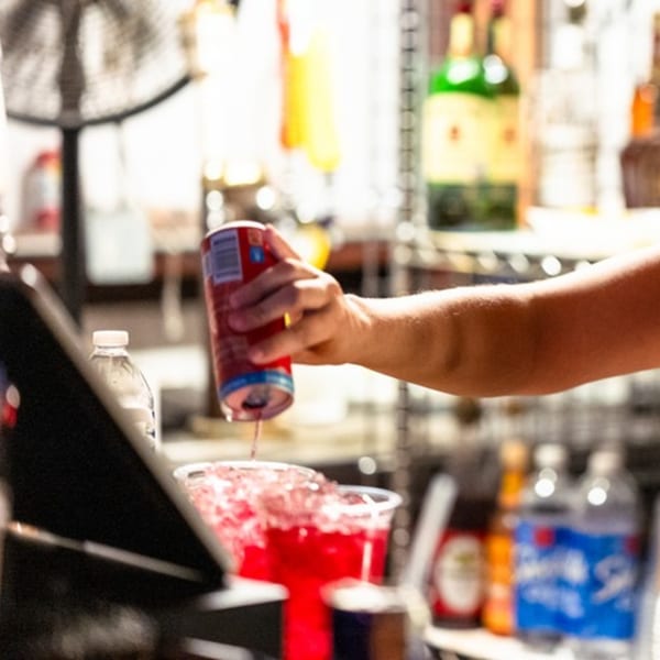 A hand pours a red canned drink into a large plastic cup filled with ice and red liquid.