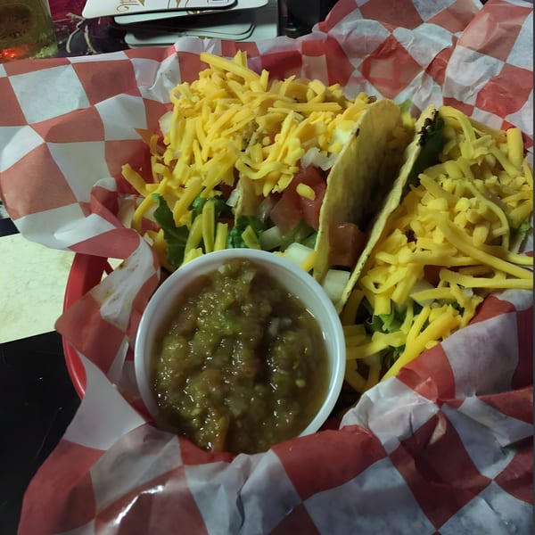 Three hard-shell tacos with shredded cheese, lettuce, and tomatoes, served with a side of salsa verde.