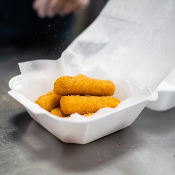 Golden-brown mozzarella sticks in a white takeout container, being seasoned by a person.