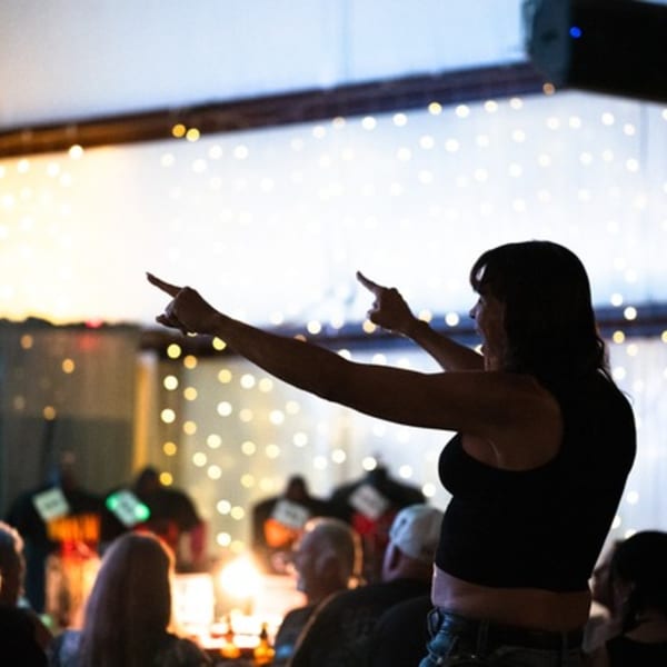 A woman in a black top points to the left and right, silhouetted against string lights.